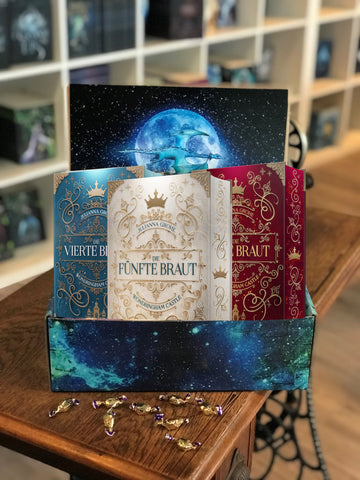 Auf dem Bild ist ein blauer Drachenpost-Karton abgebildet, welcher auf einem braunen Tisch steht. In der Box sind die drei Schmuckausgaben der Wondringham Castle-Reihe abgebildet.